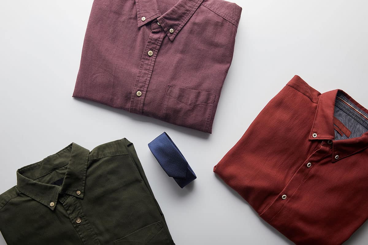 Assortment of Shirts Three neatly folded San Marcos custom t-shirts in different colors: a maroon shirt, a burnt orange shirt, and an olive green shirt, accompanied by a navy blue tie, all arranged on a light gray background.